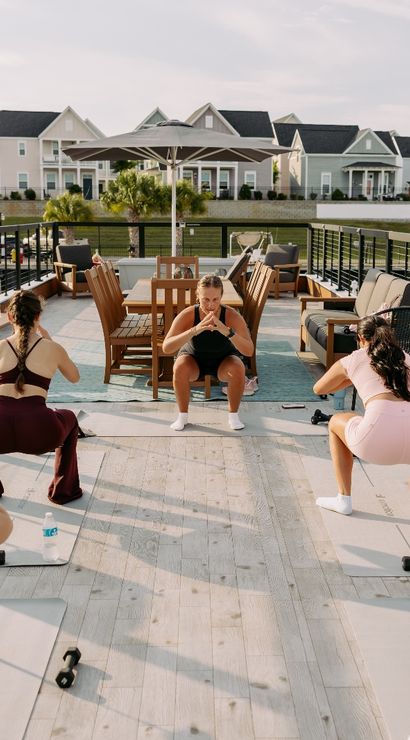Women doing a group outdoor workout on a sunlit marina deck — instructor leads squats on yoga mats with dumbbells, boats docked nearby and pastel coastal houses across the water.