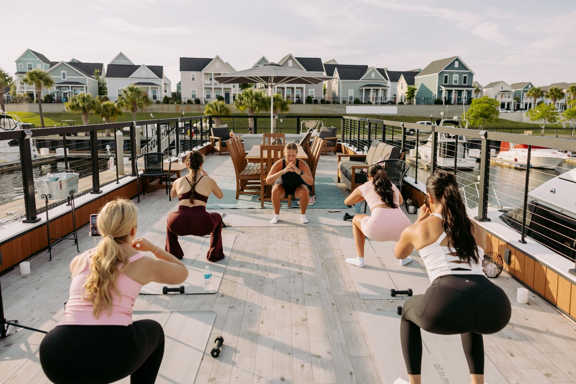 Women doing a group outdoor workout on a sunlit marina deck — instructor leads squats on yoga mats with dumbbells, boats docked nearby and pastel coastal houses across the water.
