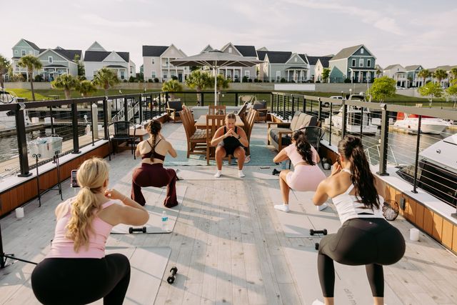 Women doing a group outdoor workout on a sunlit marina deck — instructor leads squats on yoga mats with dumbbells, boats docked nearby and pastel coastal houses across the water.