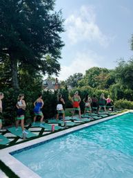 Sunny backyard poolside group fitness class — women on yoga mats doing resistance-band squats beside a turquoise swimming pool with trees and suburban houses in the background.
