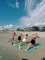 Four people doing a beach yoga class on green mats at an oceanfront shoreline, with coastal vacation homes and a bright blue sky streaked with wispy clouds.