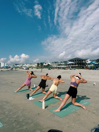 Four people doing a beach yoga class on green mats at an oceanfront shoreline, with coastal vacation homes and a bright blue sky streaked with wispy clouds.