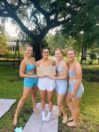 Four women in pastel blue workout sets smiling on yoga mats in a sunny suburban backyard under a large oak tree, holding a small letter board that reads “Bachelorette Pilates.”