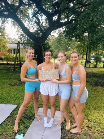 Four women in pastel blue workout sets smiling on yoga mats in a sunny suburban backyard under a large oak tree, holding a small letter board that reads “Bachelorette Pilates.”