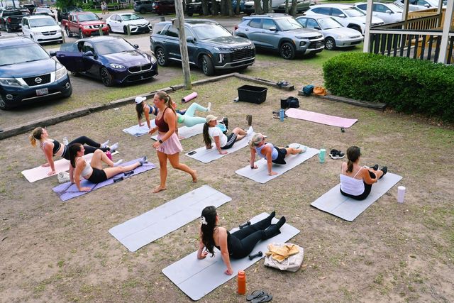 Outdoor group fitness class on grass by a suburban parking lot — women on yoga mats doing core and stretch exercises with an instructor walking among them, parked cars and shrubs in the background.