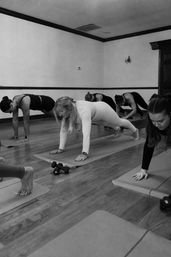 Group fitness class planking on yoga mats in a wood-floored studio, small dumbbells beside several mats.