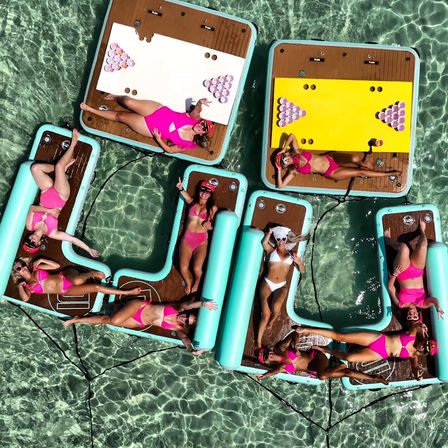Aerial view of friends in bright pink and white bikinis lounging and sunbathing on linked turquoise inflatable platforms and beer-pong-style floating boards in crystal-clear tropical shallow water