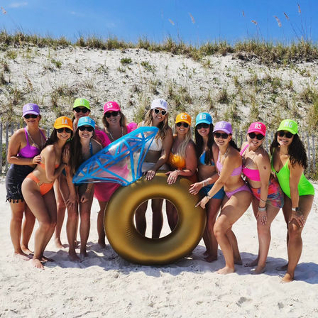 Group of women in colorful swimsuits and matching initialed caps posing on a sunny sandy beach by grassy dunes, holding an oversized gold ring float topped with a blue inflatable “diamond.”