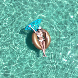 Aerial view of a person in a white bikini and cap lounging on a gold inflatable ring, waving beside a blue float in crystal-clear turquoise water — sunny tropical summer vibe.