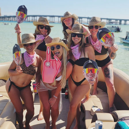Seven women in bikinis on a pontoon boat wearing straw hats and pink bandanas, holding face-cutout masks and a playful foam hand, posing on a sunny clear blue bay with a bridge in the background.