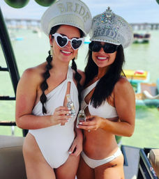 Two women in white swimsuits wearing jeweled “BRIDE” caps and sunglasses, smiling and toasting with mini liquor bottles aboard a sunny boat near a waterfront bridge and anchored float toys.