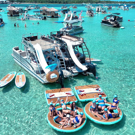 People lounging on round floating platforms beside a double-deck pontoon boat with twin water slides in crystal turquoise shallow water, surrounded by paddleboards and other boats on a sunny day.