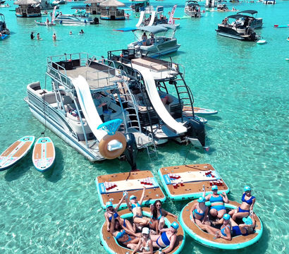 People lounging on round floating platforms beside a double-deck pontoon boat with twin water slides in crystal turquoise shallow water, surrounded by paddleboards and other boats on a sunny day.