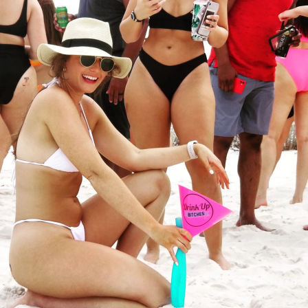 Smiling woman in a white bikini and straw sunhat kneeling on a sandy beach holding a pink "Drink Up" pennant, surrounded by friends in swimsuits with canned drinks for a lively beach party scene
