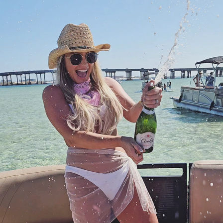 Woman in straw hat and sunglasses pops a champagne bottle on a pontoon boat in clear turquoise bay near a bridge, spraying bubbly at a sunny summer boat party