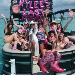 Group of women on a pontoon boat in clear turquoise water celebrating a bachelorette with pink "Last Rodeo" foil balloons, pink bandanas, oversized sparkly cowboy boot inflatables and face-cutout props, all smiling and posing in swimsuits.