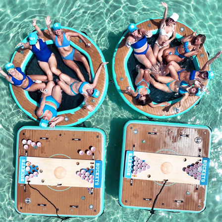Aerial drone view of a lively summer pool party: two circular floating lounges with people in blue swimwear waving and splashing in clear turquoise water, tethered to rectangular floating platforms set up with cups for a beer‑pong–style game.