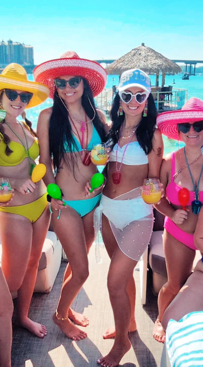 Six women in colorful bikinis and wide-brimmed sun hats on a sunny boat party over turquoise tropical water, holding cocktails and maracas with thatched tiki huts and a bridge in the background.