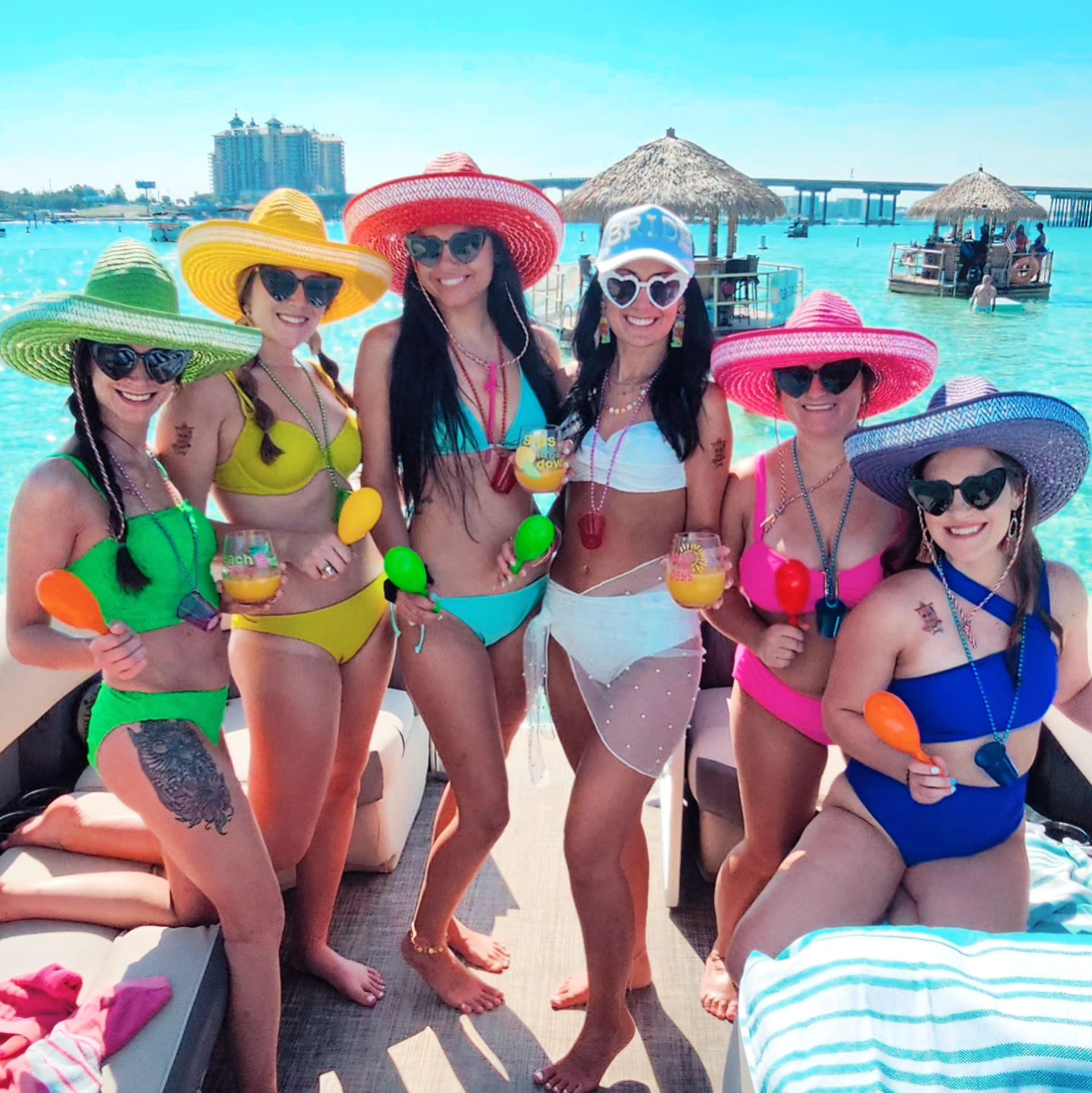 Six women in colorful bikinis and wide-brimmed sun hats on a sunny boat party over turquoise tropical water, holding cocktails and maracas with thatched tiki huts and a bridge in the background.