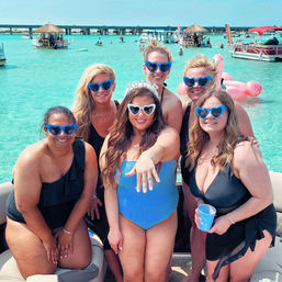 Bachelorette group on a boat in clear turquoise water — bride wearing a tiara shows her ring while friends in blue heart sunglasses smile, flamingo float and tiki huts in background