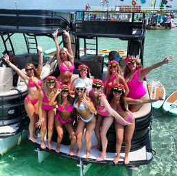 Group of women in pink bikinis and captain hats cheering on the stern of a black pontoon boat in clear turquoise shallow water, with paddleboards and a festive floating dock in the background, sunny summer boat party.