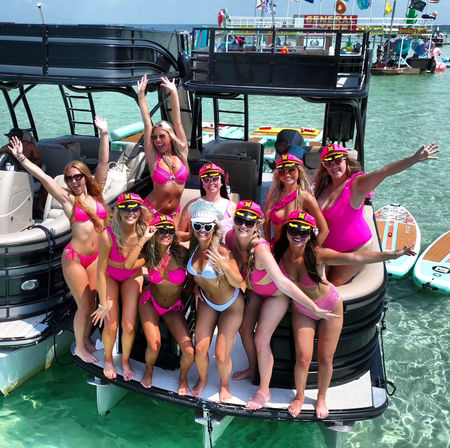 Group of women in pink bikinis and captain hats cheering on the stern of a black pontoon boat in clear turquoise shallow water, with paddleboards and a festive floating dock in the background, sunny summer boat party.