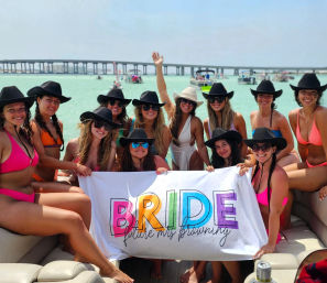 Bachelorette party on a pontoon boat in clear turquoise water with a bridge in the background — a group of women in bikinis and cowboy hats smiling and holding a colorful banner that reads BRIDE while other boats float nearby.