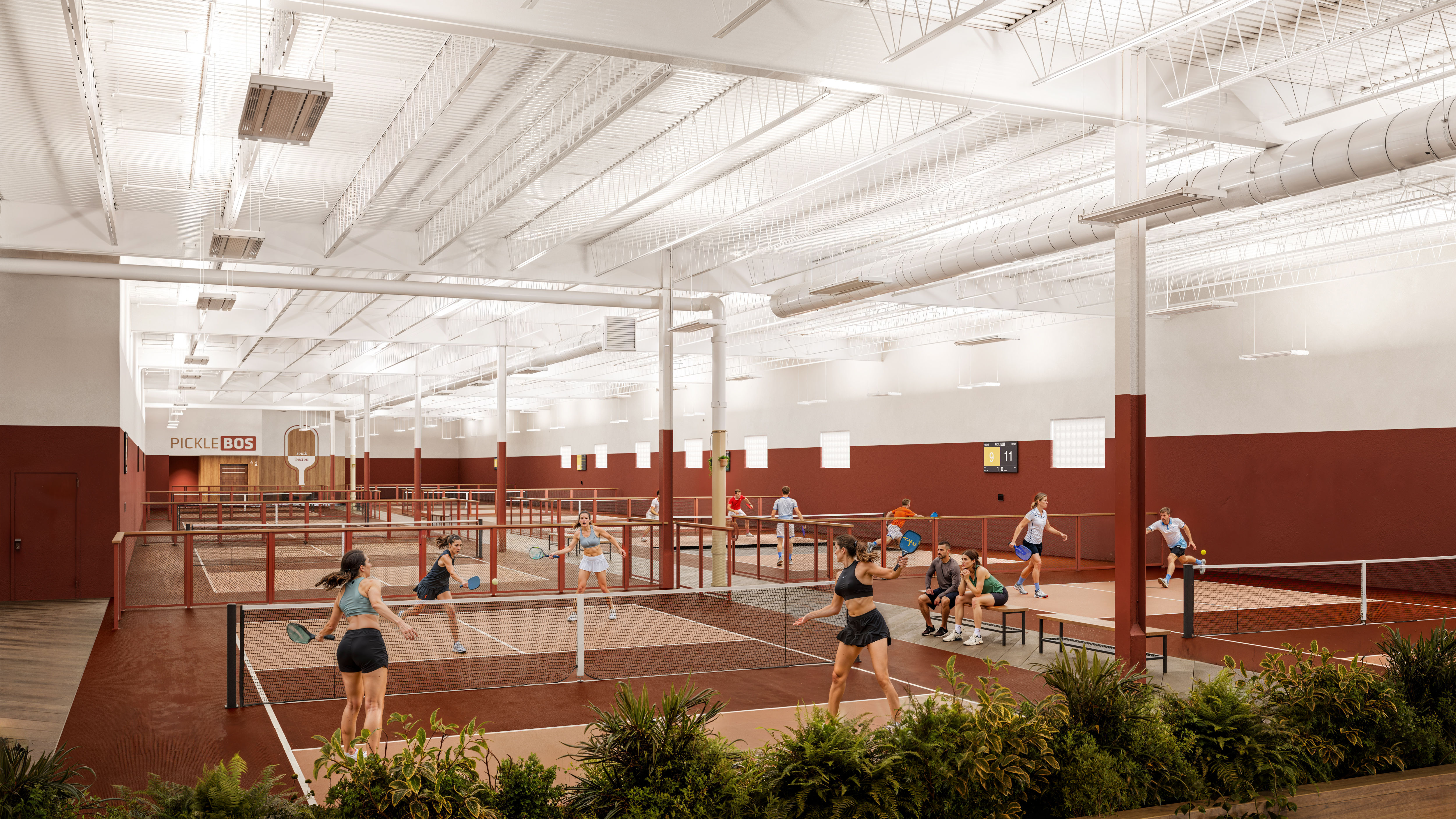 Wide-angle view of a modern indoor pickleball facility with multiple red-and-white courts, high exposed ceiling and ducts, players in action with paddles, benches with spectators, and plants in the foreground.