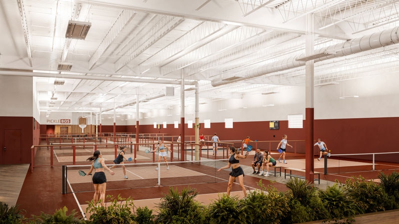 Wide-angle view of a modern indoor pickleball facility with multiple red-and-white courts, high exposed ceiling and ducts, players in action with paddles, benches with spectators, and plants in the foreground.