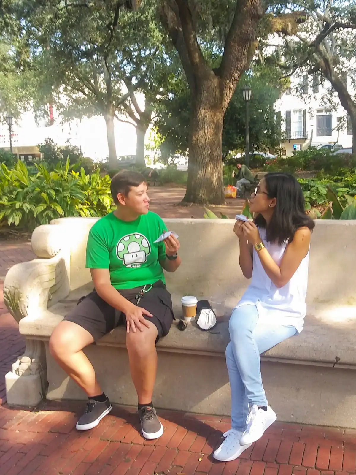 Two people sitting on a stone bench in a sunny urban park with large shade trees and brick paths, chatting while eating handheld snacks with a coffee cup on the bench.