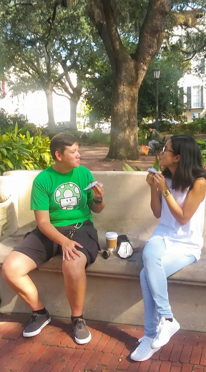Two people sitting on a stone bench in a sunny urban park with large shade trees and brick paths, chatting while eating handheld snacks with a coffee cup on the bench.