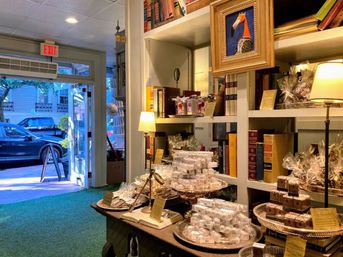 Quaint boutique shop interior with bookshelves, framed art and warm lamps, tables stacked with wrapped artisan chocolates and confections on silver trays, and an open glass door showing the street and parked cars.