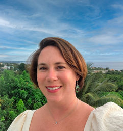 Smiling woman selfie on a balcony overlooking lush tropical greenery and a calm coastal ocean under a blue sky