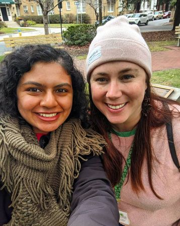 Outdoor selfie of two smiling women bundled for winter—one in a chunky knit scarf, the other in a pink beanie—sitting on a park bench in a tree-lined residential neighborhood with brick sidewalk and parked cars.