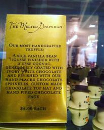 Gold menu card describing a handcrafted vanilla-bean mousse truffle finished with cognac and ivory white chocolate, beside a glass display of snowman-shaped white chocolate truffles with chocolate top hats — $4 each.