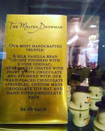 Gold menu card describing a handcrafted vanilla-bean mousse truffle finished with cognac and ivory white chocolate, beside a glass display of snowman-shaped white chocolate truffles with chocolate top hats — $4 each.