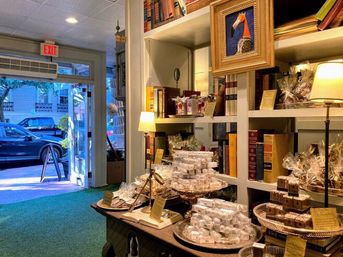 Cozy boutique gift shop interior with stacks of gourmet chocolates and wrapped confections on a wooden table, books and warm lamps on shelves, framed art, and an open door to a tree-lined downtown street.