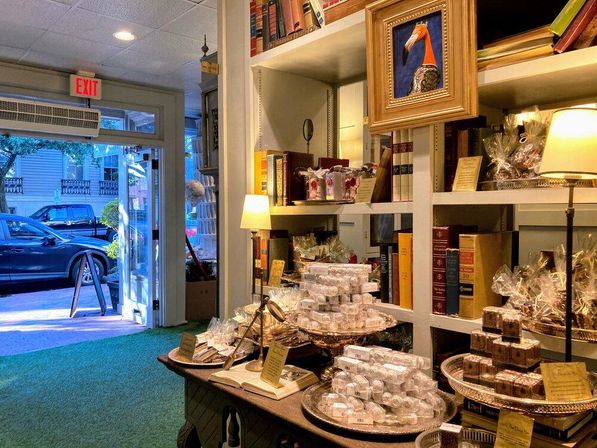 Cozy boutique gift shop interior with stacks of gourmet chocolates and wrapped confections on a wooden table, books and warm lamps on shelves, framed art, and an open door to a tree-lined downtown street.
