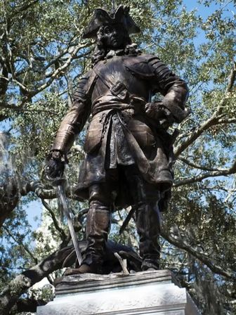 Bronze statue of a colonial-era soldier in a tricorn hat holding a sword, standing on a stone pedestal beneath moss-draped oak trees and a blue sky.