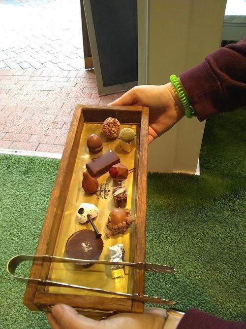 Tempting assortment of gourmet chocolates and truffles arranged on a gold‑lined wooden serving tray with metal tongs, hands holding it over green turf by a shop doorway.