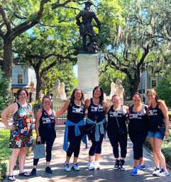 Seven friends in matching tank tops pose before a bronze statue in a sunlit southern park with Spanish-moss draped live oak trees.