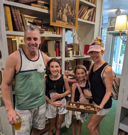Smiling family of four in casual summer clothes holding drinks and a wooden tray of mini desserts inside a cozy bookstore with shelves of vintage books.