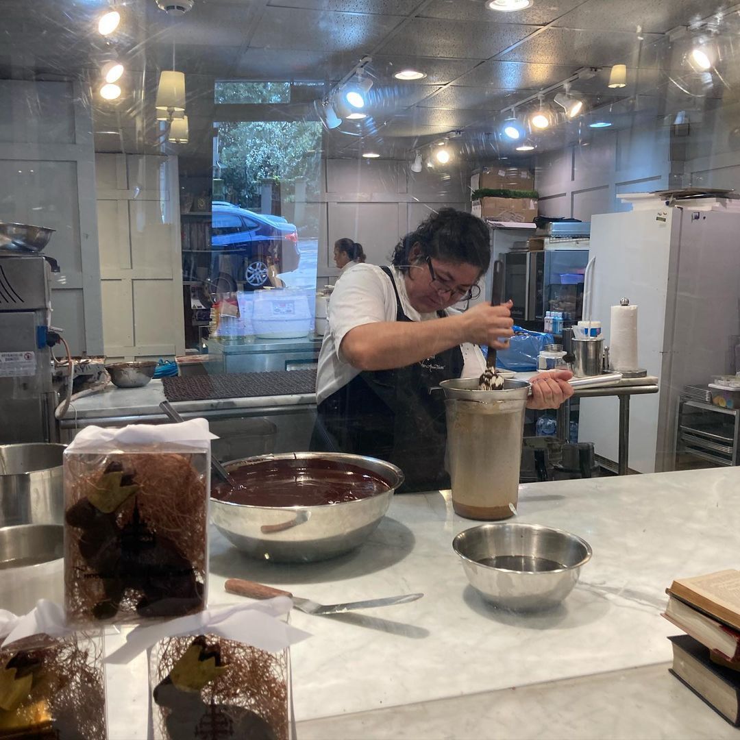 Artisan chocolatier at work behind a shop window, stirring melted chocolate in a tall tempering tank with metal bowls and packaged truffles displayed on a marble counter in a modern pastry kitchen.