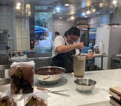 Artisan chocolatier at work behind a shop window, stirring melted chocolate in a tall tempering tank with metal bowls and packaged truffles displayed on a marble counter in a modern pastry kitchen.