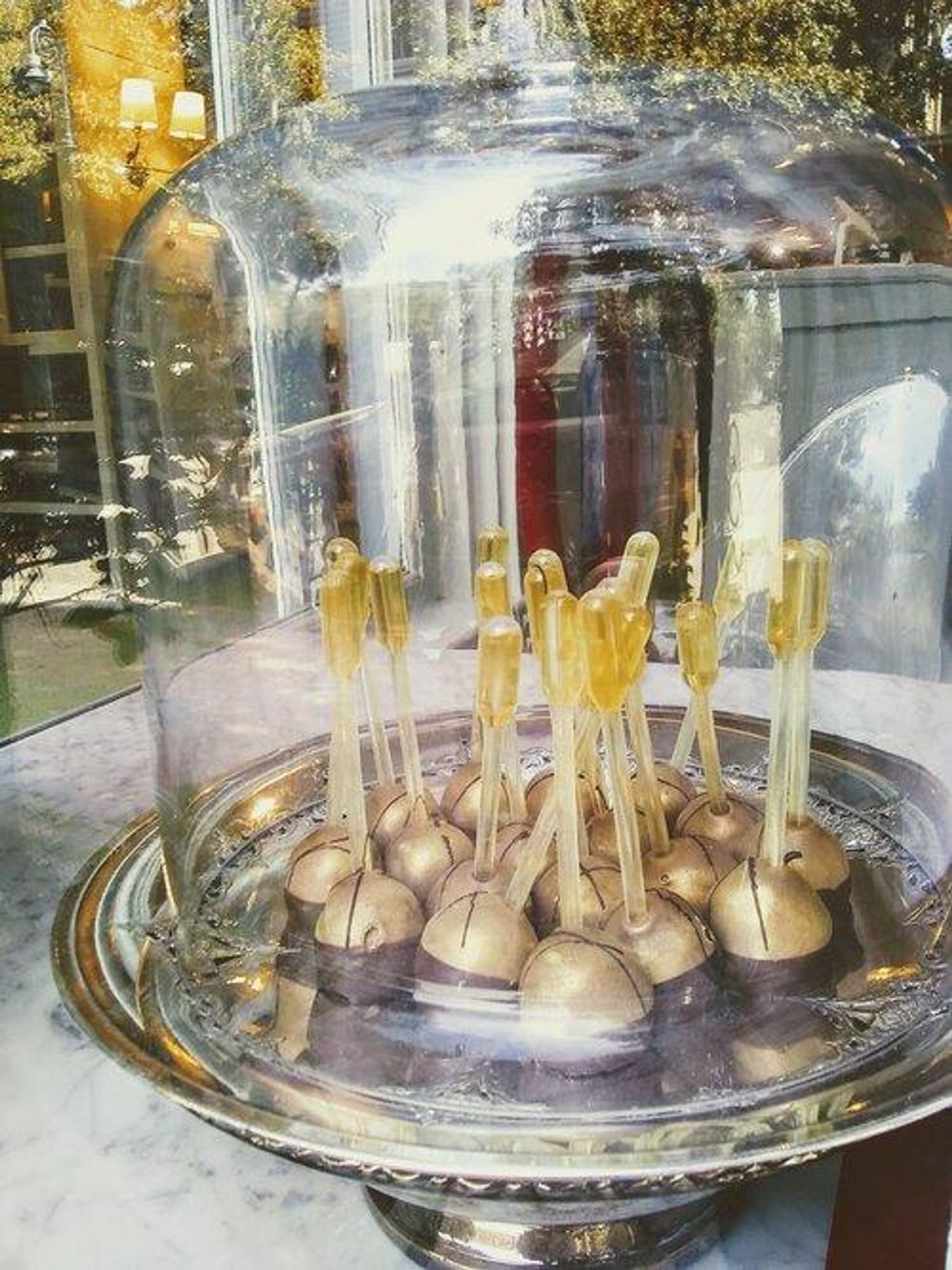 Gleaming gold-topped cake pops on clear sticks arranged on a silver platter under a glass cloche in a bakery window display, with street reflections and warm interior lamps