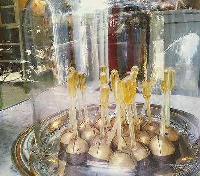 Shiny gold-coated cake pops on clear sticks arranged on an ornate silver platter under a glass cloche in a bakery window with street and lamp reflections.