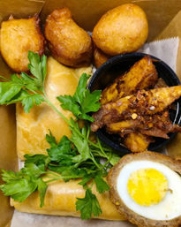 British-style takeaway snack box with halved scotch egg showing bright yolk, golden sausage rolls, crispy fried dough balls, spicy seasoned wedges in a black cup and fresh parsley garnish