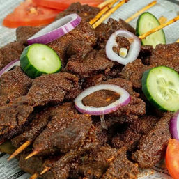 Close-up of spicy West African-style grilled beef skewers (suya) piled on skewers, garnished with cucumber slices, red onion rings and tomato wedges