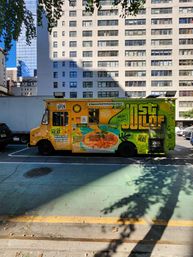 Vibrant yellow-green food truck with a large illustrated bowl of rice and meat and an 'OPEN' sign, parked on a sunny urban street curb in front of a tall apartment building.