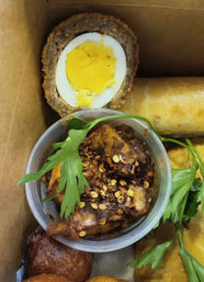 Takeout box of appetizers: halved scotch egg, crispy spring roll, and a cup of caramelized fried plantain bites sprinkled with red chili flakes and parsley garnish.